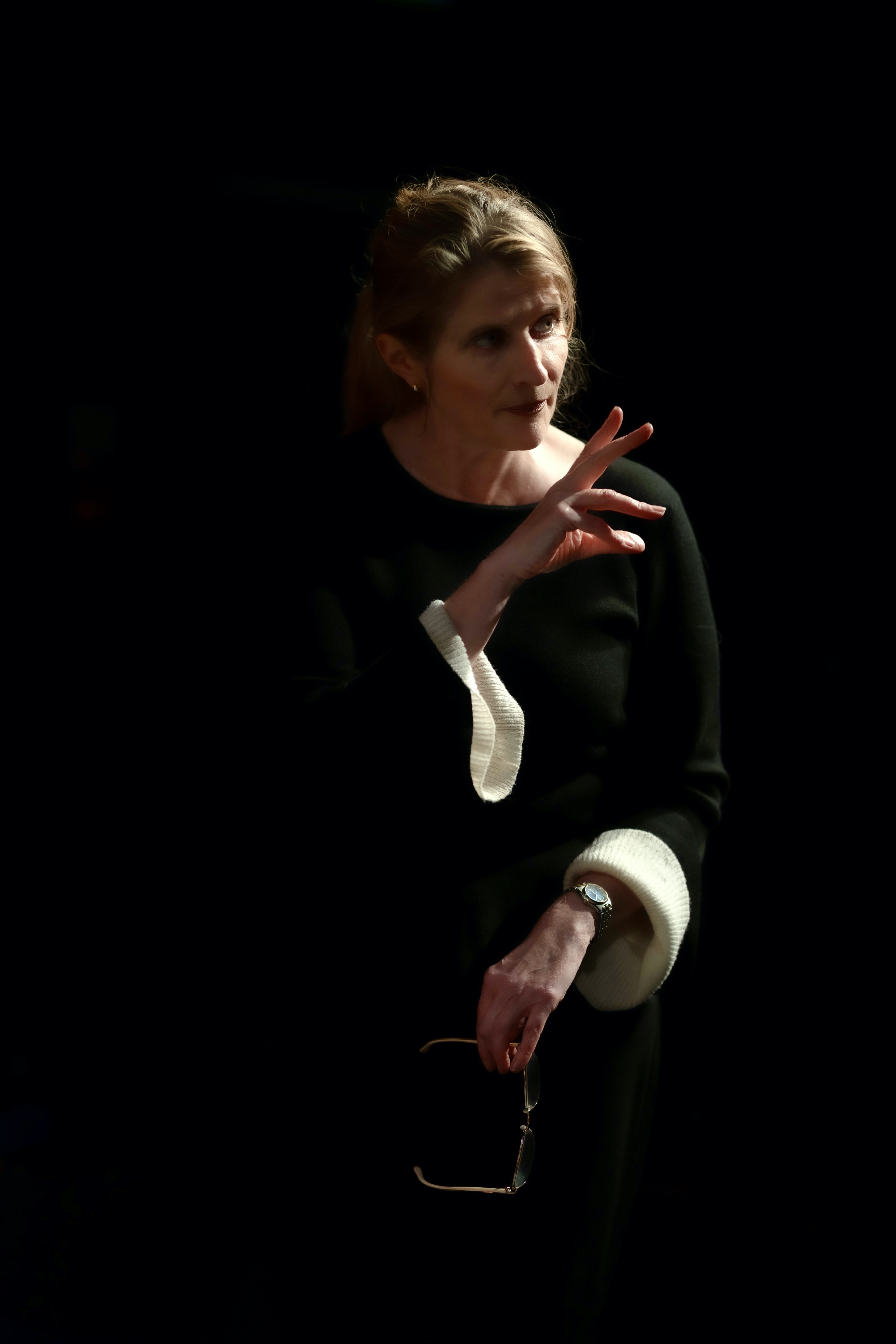 Jude Haste, a white woman lit against a black background, making an expressive gesture with her fingers.