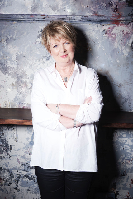 Fiona Talkington, a white woman with cropped short blond hair, smiling with her arms crossed standing against a metal background.